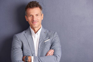 Close-up portrait shot of confident middle aged businessman with folded arms standing at grey wall and looking at camera.