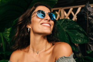 Portrait of cheerful tanned young ethnic brunette in sunglasses smiling and looking away while spending summer day in green tropical park