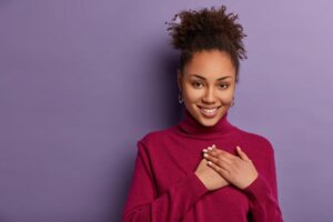 Smiling lovely curly girl impressed by touching moment, presses palms to chest, feels thankful for help, has curly combed hair, wears casual burgundy jumper, models over purple wall, blank space