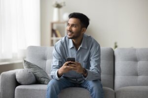 Positive pensive freelance Indian business man enjoying online communication at home, holding smartphone, looking at window away, smiling, thinking, using Internet technology on gadget for work